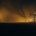 Una torre de piedra aislada a la izquierda y un entramado de estructuras industriales iluminadas a la derecha, separadas por una llanura a oscuras, a contraluz ámbar