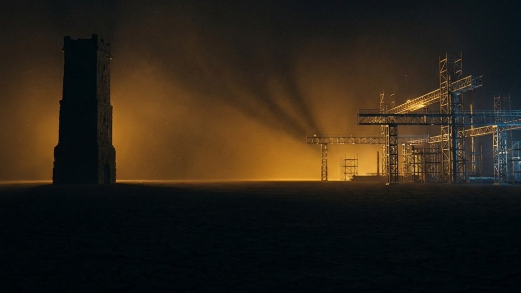 Una torre di pietra isolata a sinistra e un intrico di strutture industriali illuminate a destra, separate da una piana al buio, in controluce ambra