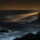A lighthouse on a cliff at night lights up a bay full of cargo ships at anchor, backlit in amber.