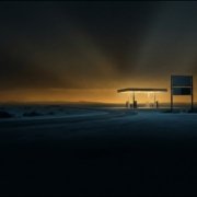 Una gasolinera aislada en una carretera desierta de noche, con la marquesina iluminada y el cartel de precios apagado, bajo un resplandor ámbar en el horizonte