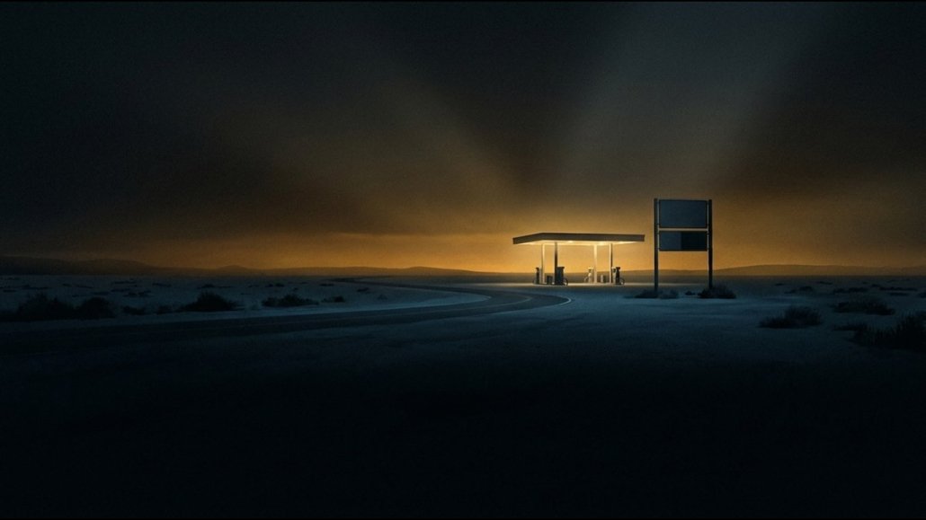 Una stazione di servizio isolata su una strada deserta di notte, la pensilina illuminata e un cartello dei prezzi spento, con un bagliore ambra all'orizzonte