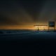Una stazione di servizio isolata su una strada deserta di notte, la pensilina illuminata e un cartello dei prezzi spento, con un bagliore ambra all'orizzonte