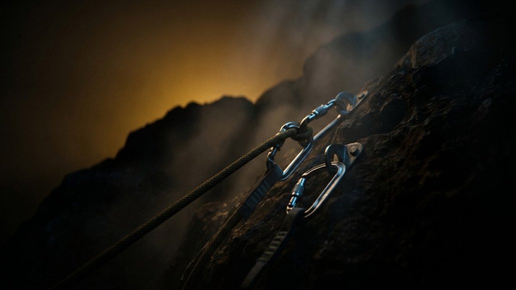 A climbing rope secured with steel carabiners to two bolts on a dark rock face, in the haze and backlit in amber.