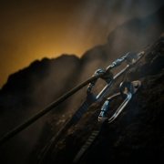 Una corda d'arrampicata assicurata con moschettoni d'acciaio a due chiodi su una parete di roccia scura, nella foschia e in controluce ambra.